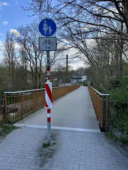 Es ist der Anfang der Brücke zu sehen, Fahrtrichtung abwärts. Der Pfosten für die Schilder "Fußweg" und "Radradfahrende frei" ist mittig platziert. Zur besseren Sichtbarkeit ist hier un eine reflektierende, circa halben Meter hohe reflektierende dünne Tonne angebracht worden.