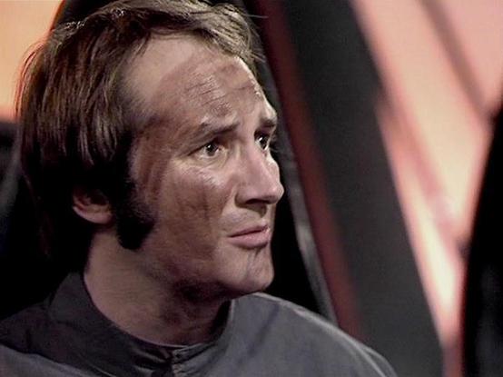 Claude 3.7 describes the image as: "This image appears to be from a science fiction television series, showing a profile view of a person in what looks like a futuristic setting. The individual is wearing what appears to be a dark, possibly uniform-like outfit. They have distinctive sideburns and a hairstyle that suggests the production is from the late 1970s or early 1980s era.

The background has a reddish-pink tint with some structural elements visible, which fits with the aesthetic of sci-fi productions from that period. The lighting and set design have that characteristic studio television production quality of that era.

The person appears to have a concerned or thoughtful expression, captured in what seems to be a dramatic moment from the show."

Blakes7Bot, version 3.9.33.