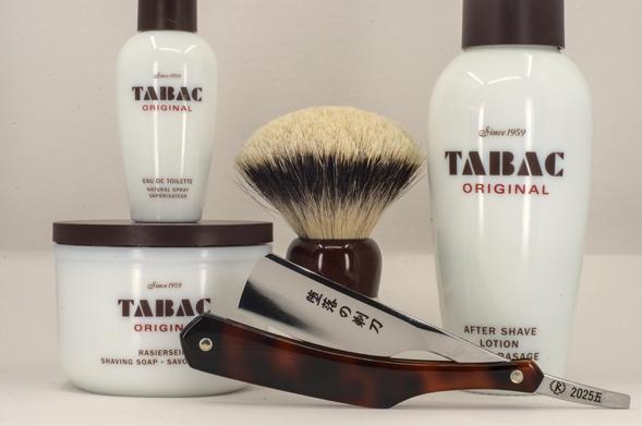 (Le Chat Mistral description)

This image shows a classic shaving setup arranged on a flat surface. On the left, there is a white container of Tabac Original shaving soap. Behind it, there is a bottle of Tabac Original eau de toilette. To the right of the soap, there is a bottle of Tabac Original aftershave lotion. In front of the shaving soap, there is a shaving brush with a dark handle and light bristles. Resting diagonally across the items is a straight razor with a tortoiseshell-patterned handle and a steel blade, which has 堕落の剃刀 (Razor of Depravity) and the marking "2025.五" etched on it. The items are neatly organised, suggesting a traditional wet-shaving experience.