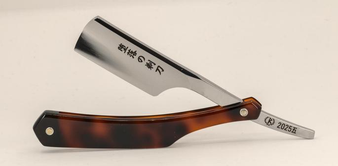 (Le Chat Mistral description)

This image shows a folded straight razor placed on a plain, light background. The razor features a polished, metallic blade with a sharp edge. The handle is made of a material that resembles tortoiseshell, with a mix of dark and light brown tones. The blade is etched with Japanese characters that read "堕落の剃刀", translating to "The Depraved Razor" or "Razor of Depravity". The razor's pivot area is metallic, and near the end of the blade, there is an inscription that includes "2025.五", indicating May 2025. The razor is elegantly designed, reflecting a blend of traditional craftsmanship and stylistic detail.