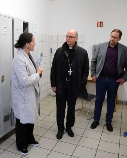 Der Bischof von Würzburg spricht in der Gemeinschaftsküche mit einer Frau in einem grauen Mantel in einem Raum mit Spinden.