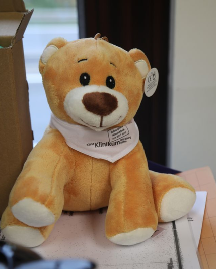 Plüschbär mit braunem Gesicht und weißen Akzenten trägt ein Halstuch mit dem Aufdruck 'Klinikum Würzburg Mitte'.