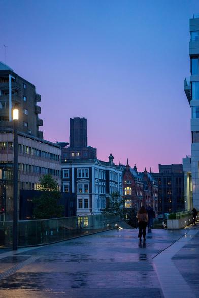This image depicts an urban scene at dawn, with a sky transitioning from blue to purple hues. In the foreground, there is a wide, reflective pedestrian walkway that appears to be slightly wet, possibly from recent rain. Two people are walking away from the camera on this walkway, one slightly ahead of the other. The walkway is bordered by a glass railing on the left side, reflecting the surrounding buildings and lights.

On the left side of the image, there are modern buildings with large windows, some of which are illuminated from within. One of the buildings has a prominent light fixture on its exterior. In the background, there is a row of older, ornate buildings with intricate architectural details, such as arched windows and decorative facades. These buildings are also lit from within, adding warmth to the scene. The overall atmosphere is calm and serene, with a mix of modern and historical architecture under a softly lit sky.