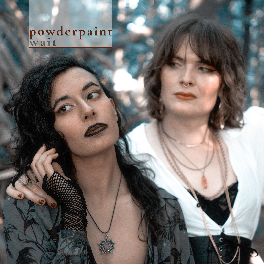 cover art for a single by powderpaint called 'wait' - shonalika, a brown goth with long black hair and dark make-up, stands in a forest looking wistfully off-camera. hazel, a white trans fem with shaggy brown hair, stands next to shonalika looking at them
