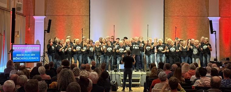 Das Bild zeigt den RockChor60+ auf der Bühne des 2. Chorfestivals Mönchengladbach.

Über 40 Stimmen formieren sich zu einem Chor. Sie tragen schwarze Oberteile mit der Aufschrift "RockChor 60+" und Blaue Jeans.