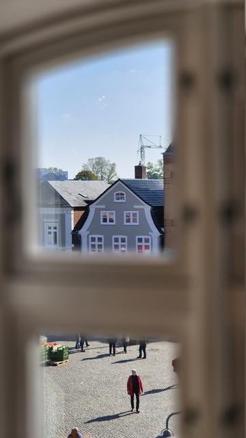 Aus einem oberen Stockwerk im Inneren, dicht vor einem Sprossenfenster, dessen Fensterrahmen durch die Nähe nur sehr unscharf zu erkennen ist, richtet sich der Blick durch zwei durch eine Quersprosse getrennte Fensterscheiben auf das Treiben vor dem Haus und die gegenüberliegenden Gebäude im historischen Stil.