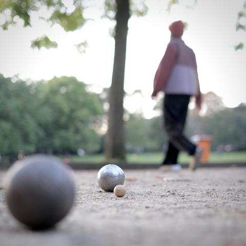 Boulekugeln im Licht der Dämmerung. Eine Frau läuft im Hintergrund durchs Feld.
