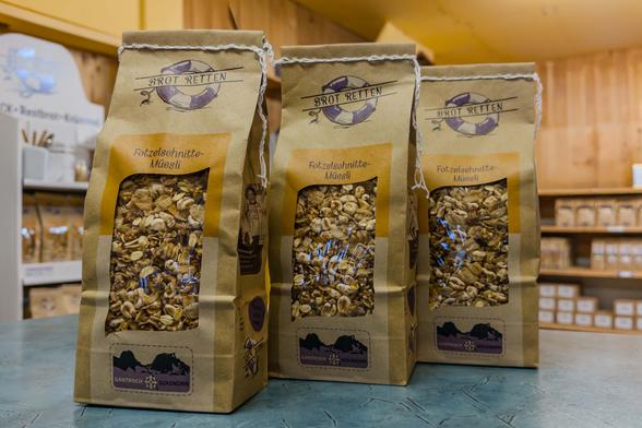 Eine Reihe von fünf braunen Papiertüten mit Sichtfenster, gefüllt mit Fotzelschnitte-Müesli (Arme Ritter oder Povesen). Auf der Packung steht das Label «Brot Retten» mit einem Rettungsring-Logo, darunter ein Cellophan-Sichtfenster, durch das man das Müesli sieht. Die Packungen stehen auf einem Holztisch, im Hintergrund sind weisse Kartons gestapelt.