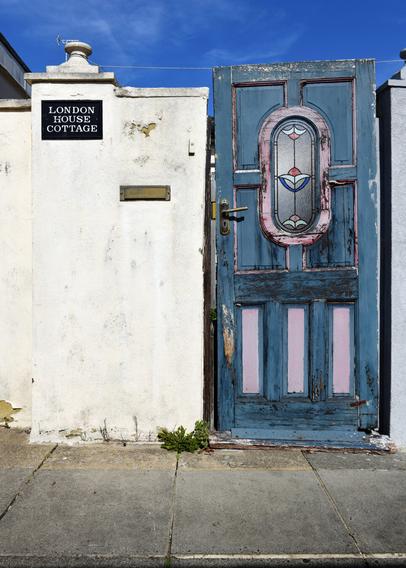 Photo of a dilapidated front door. A panel door with coloured glass insert, originally painted blue and pink but now very faded and peeling.