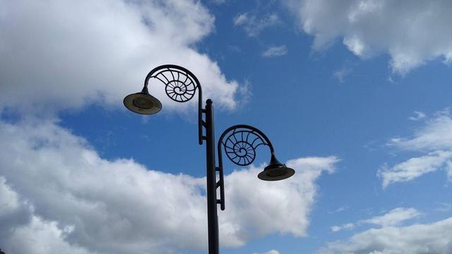 A lighting column with two flat conical lanterns. The bracket is a curved arch into which there is an outline of an ammonite curling into the space under the arch. The assembly is black and viewed against a blue sky with white fluffy clouds.