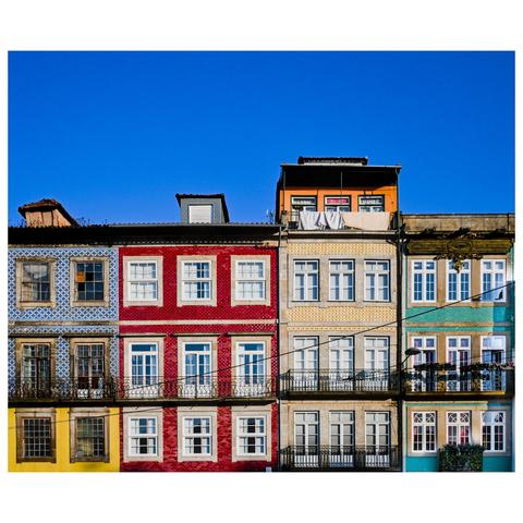 This image showcases a row of colorful, traditional buildings in Porto, Portugal. Each building features a distinct facade, painted in vibrant colors such as blue, red, yellow, and green. The facades are adorned with decorative tile patterns, particularly the blue and white azulejos, which are characteristic of Portuguese architecture. The buildings have multiple windows with white frames, wrought-iron balconies, and some have laundry hanging out to dry, adding a touch of everyday life to the scene. The clear blue sky in the background enhances the vividness of the colors and the overall charm of this picturesque urban landscape. (created with mistral.ai)