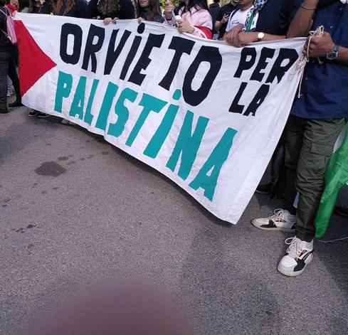 Delle persone, di cui si vedono braccia, petti e spalle, reggono uno striscione sul quale è scritto, in nero e verde, Orvieto per la Palestina. Sulla sinistra dello striscione è disegnato un triangolo rosso come nella bandiera palestinese
