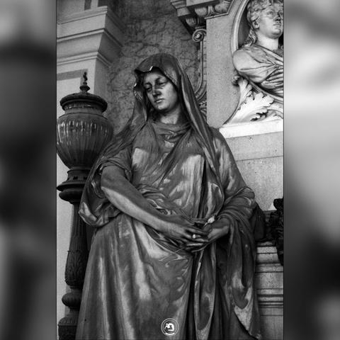 Black and white photo of a grave ornament/statue. The statue is a hooded woman, looking off to the side of the photo. She is leaning against the tomb, cradling her hands in each other.