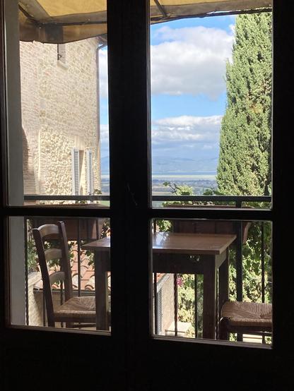 View through a tall window over a balcony with a table outside. In the distance is a lake and hills beyond.  A stone building is in the left side of the frame and a tall cedar fills the right side of the frame.
