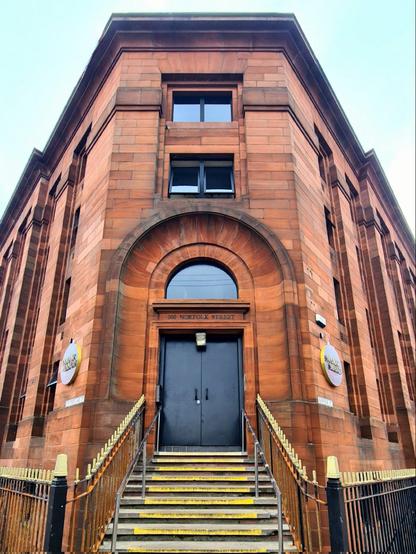 A Romam-arched doorway at the top of a flight of steps in a red sandstone former library building.