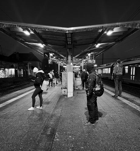 DE: Schwarzweiß-Foto eines überdachten Bahnsteigs längs gesehen. Es ist dunkel und auf ihm stehen, locker verteilt, im Schein der Leuchten Menschen in winterlicher Kleidung. Der Zustand wirkt etwas heruntergekommen EN: Black-and-white photo of a covered platform seen lengthwise. It is dark, and people in winter clothing are scattered loosely across it, illuminated by the lights. The condition appears somewhat dilapidated.