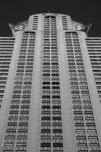 This is a black and white photo of the Art Deco Chrysler Building on Lexington Ave in New York City (2012).
The camera is tilted upwards at a steep angle towards the top of this historic and iconic Manhattan high-rise. The image is composed around the centre of the Lexington Ave facade which has rendered to a medium grey or silver tone made up of masonry tiles or bricks and rectangular windows with highly reflective metal being used decoratively towards the top. The building is slightly tiered, the lower floors being broader than those higher up. There are six columns of windows in view, arranged in pairs with a wide vertical strip of masonry dividing each pair. There are about thirty-five floors in view in the centre (out of a total of seventy-seven) with less in the outer tiers of the building. Towards the top, the masonry arches over forming a semicircle with the stainless steel forming multiple arches above, each one smaller than the one below, forming a spire - the spike on the top being just in view. In two if the corners of the centre square section of the building, on the sixty-first floor, are two large and ornate stainless steel, Gargoyle like Eagles that protrude outwards  overhanging the building by a few metres. 
Above is a clear and cloudless sky, rendered jet black.