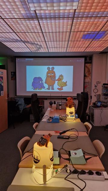 Das Bild zeigt einen hell beleuchteten Raum mit einem Präsentationsbildschirm, der animierte Charaktere zeigt: einen Elefanten, eine Maus und eine Ente. Im Vordergrund ist ein Tisch mit einer Lampe mit einem Stofftier, zusammen mit verschiedenen Werkzeugen und Materialien zu sehen.