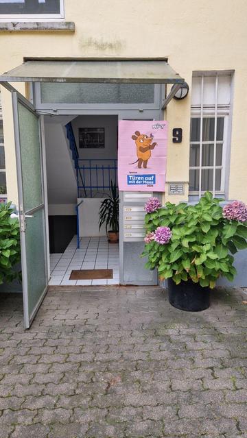 Ein Eingang mit einer Glastür und einer sichtbaren Treppe, die in die oberen Etagen führt. Neben der Tür ist ein großes Poster mit der Maus zu sehen, das für eine Veranstaltung wirbt. Es gibt Briefkästen neben dem Eingang und in der Nähe blüht eine Pflanze.