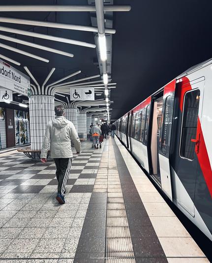 DE: Foto des Bahnsteigs einer unterirdischen U-Bahnstation der Hamburger Hochbahn, rechts wartet ein Zug mit offenen Türen, die sternförmig angeordneten Dekoelemente steht in starkem Kontrast zur schwarzen Decke und lässt die Station höher wirken. Die Farben weiß und shwarz dominieren die Szene und stehen im starken Kontrast zur einer roten Linie als Dekoration am Zug. Die Menschen in alltäglicher, herbstlicher Kleidung auf dem Bahnsteig bewegen sich vom Fotografen weg. EN: Photo of the platform of an underground subway station of the Hamburg Hochbahn. On the right, a train waits with its doors open. The star-shaped decorative elements contrast sharply with the black ceiling and make the station appear higher. The colors white and black dominate the scene and contrast sharply with a red line used as decoration on the train. The people on the platform, dressed in everyday autumn clothing, are moving away from the photographer.