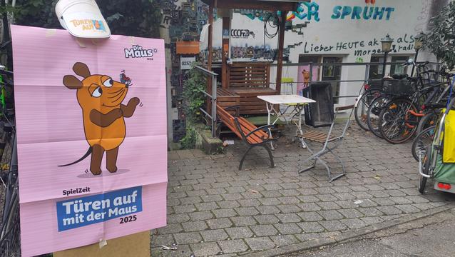 Ein grosses rosa Plakat mit der Maus und 'Türen auf mit der Maus 2025', im Hintergrund ist der Eingangsbereich des CCCFr zu sehen. Auf der Fassade kleben alte Platinen.