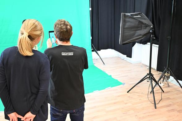 Foto in einer Fotostudio-Situation. ein Mensch hält ein Tablet, ein zweiter Mensch schaut dabei zu. vor einem Greenscreen steht eine kleine Person (von der zweiten Person fast verdeckt). Rechts eine Fotolampe.