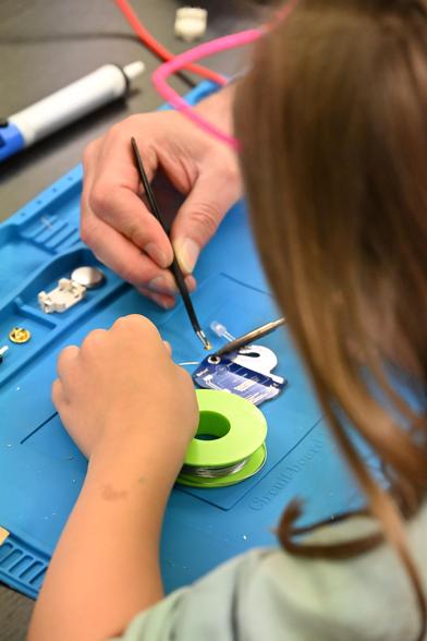 einer jungen Person über die Schulter geschaut, die lötet. Auf einer blauen Lötunterlage liegt eine lilafarbene Platine, Lötkolben, Lötziin, eine Hand einer erwachsenen Person hält eine Pinzette.