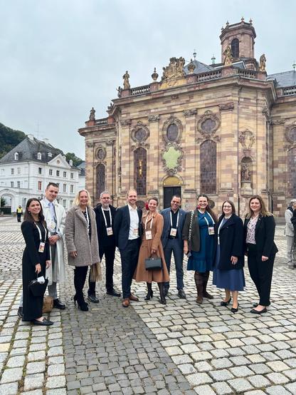Gruppenbild mit festlich gekleideten Menschen auf einem gepflasterten Platz vor einer barocken Kirche