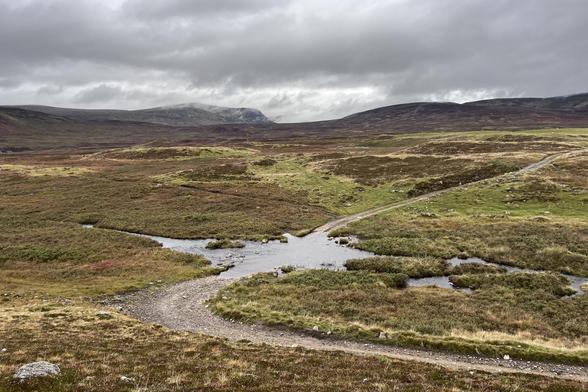A heather strewn rolling highland moorland on a grey day. A track snakes its way through the landscape, blocked by a flooded ford from a normally small stream in the foreground. In the distance is the dark peak of A’Chailleach, part of the Monadhliath mountain range.