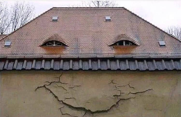 A beige house wall with a red roof. There are two windows with round arches in the roof. In the wall directly below, there's a crack that forms a downward-pointing arch. It looks like a slightly stoned smiley face.
