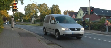 Ein PKW ist über eine ampelgeregelte Querungshilfe gefahren. Die Ampel zeigt bereits orange. Ist allerdings gerade erst umgesprungen, als das Fahrzeug bereits gequert hatte. Auf den Gehwegen warten Menschen.
