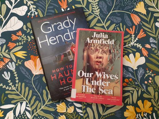 Kuva kahdesta kirjasta kuviollista tapettia vasten. Kirjat Grady Hendrixin How to sell a haunted house ja Julia Armfieldin Our Wives under the sea