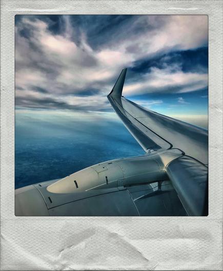 A color photo showing a view of a landscape from an airplane window. Part of the airplane wing and engine are visible in the lower section of the frame. The sky is dramatically cloudy stretching to the horizon. Framed in white.