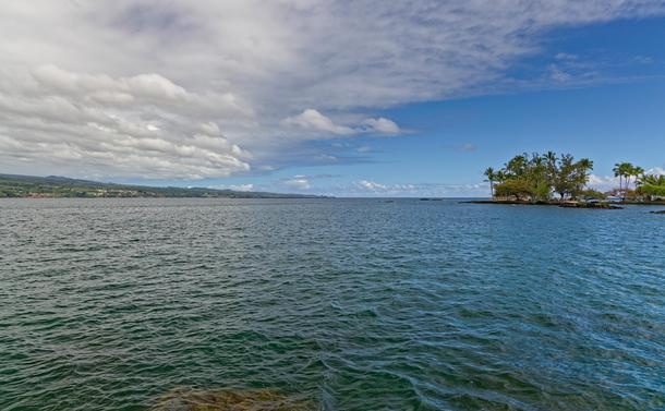 A landscape picture of Hilo bay with the east coast of the island to the left of the shot and a small island with trees to the right. The bay is a blue-green color and grey clouds fill the sky to the left drifting to center.