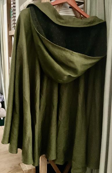 Ein Cape nit Kaputze hängt auf einem Kleiderbügel. Rechts ein Vorhang unden und Links ein Eichentisch