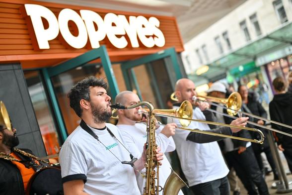 Musicians playing saxophone and trombones in a New Orleans‑style brass band outside Popeyes Swansea during the opening celebrations.