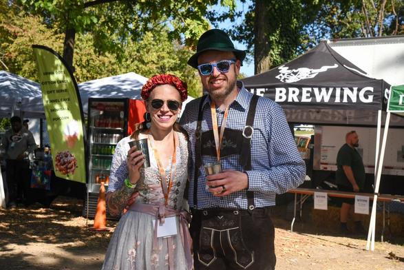 In the image, two people, a man and a woman, are standing close together and smiling, possibly posing for a photo. Both individuals are wearing colorful clothing and holding cups. The man is holding a cup in his left hand, while the woman has her right hand tucked into her pocket.

In the background, there are two tents visible, possibly indicating a festival or outdoor event. These tents are situated on the left side of the image, with one closer to the center and the other slightly further back. Additionally, there is a car parked in the vicinity of the tents.