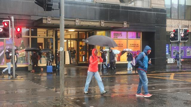 Lady in bright red coat with her hood up, sheltering under an umbrella while crossing a rainy street