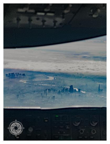 The skyscrapers of London rise over the mist with a cloud bank in the distance. The river Thames snakes from right to left as it glistens in the early morning sun. The overhead panel of a Boeing 787 and the mode control panel frame the top and bottom of the picture, cast in shadow against the daylight.
