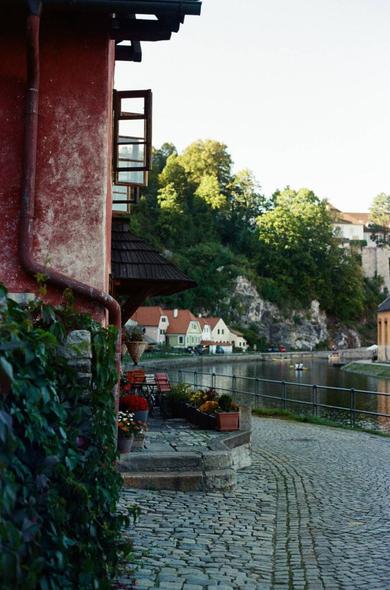 A photograph of a peaceful riverside scene in Český Krumlov, Czech Republic. On the left side of the image, part of a rustic red building with an open wooden window is visible, its wall textured with age. Beneath it, a cobblestone path curves gently along the riverbank. Small tables, chairs, and flower pots filled with colorful blossoms line the walkway, suggesting a quiet café or terrace. The river, calm and reflective, runs through the center of the image. On the opposite side of the river, pastel-colored houses with red roofs sit at the base of a steep rocky hillside covered in trees. A few people can be seen in the distance, enjoying a boat ride on the water. The overall atmosphere is tranquil, with soft evening light illuminating the scene, evoking the timeless charm of a medieval riverside town.