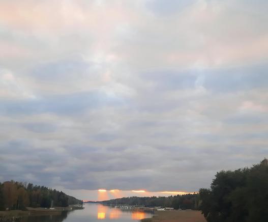 Abendrot mit durchbrechenden orangefarbenen Sonnenstrahlen unter einer dichten Wolkendecke. Links und rechts bewaldetes Land, mittig ein Ostsee-Arm, vorne rechts etwas Strand, im Huntergrund rechts ein kleiner privater Schiffsanlegeplatz.