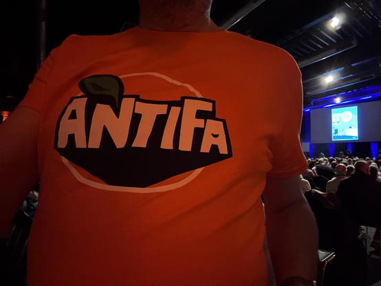 Person in einem orangenen T-Shirt mit der Aufschrift ‘ANTIFA’ in stilisierter Schrift, die an ein Orangen-Logo erinnert. Im Hintergrund sitzen viele Menschen in einem Saal, vorne ist eine große Leinwand mit einem Ruthe-Cartoon zu sehen.
