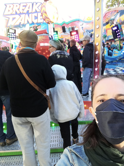 a masked selfie of a white woman standing in front of a fair ride called Break Dance. Various other people are milling about.