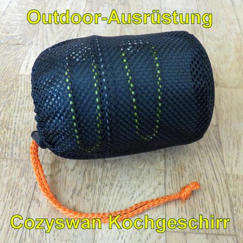 Ein kompaktes Outdoor-Kochgeschirrset, verpackt in einem schwarzen Netzbeutel mit einem orangefarbenen Kordelzug. Das Geschirr ist durch das Netz teilweise sichtbar, mit grünen Akzenten. Die Form ist zylindrisch und wirkt leicht und transportabel, ideal für Camping oder Wanderungen.