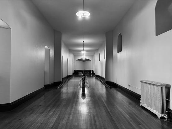 Black and white image of an underground indoor bowling alley in the Biltmore House. The view is straight down the alley with the ball return in the middle. The floor is polished wood. There are hanging lights also down the center of the alley. The walls are plain white, and there is a radiator for heat on the right wall. Biltmore House, Asheville, North Carolina.