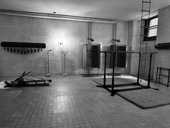 A black and white image of an underground gum room that is covered with tile. Various old gym equipment is in the room: parallel bars, mats, rope ladder, perhaps a rowing machine, scales, bowling pins, and two small showers. There is one small window to the right. Biltmore House, Asheville, North Carolina