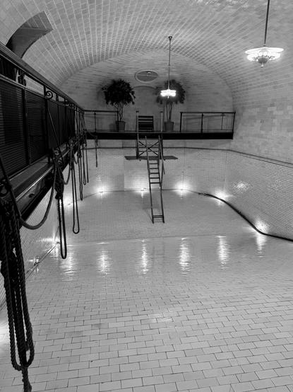 Black and white image. We are standing at the shallow end of an empty indoor swimming pool, looking towards the deep end. The pool and the room are all white tile. There are a couple of hanging lights over the pool, as well as what would be underwater lights around the bottom of the pool. There is a balcony/walkway on the left that has a metal rail as well as ropes hanging from the rail. At the far end, the balcony/platform continues and a ladder goes down to the bottom of the deep end. There was probably a diving board in this location. Two potted trees are also on the platform. Biltmore House, Asheville, North Carolina.