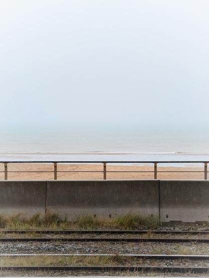 Blick von den Gleisen der Küstentram über Betonmauer und Geländer hinweg auf den Strand und die Nordsee, die im Regen und Nebel kaum vom Himmel zu unterscheiden ist. Foto by Johann Seidl, Gartenpoet.