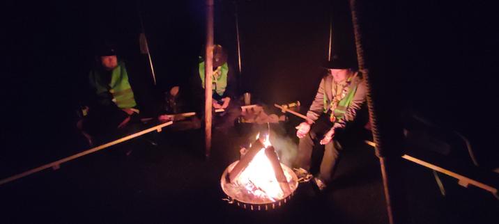 Lagerfeuer in Pfadfinderjurte auf Biertischbänken sitzen die 3 Pfadfinder der Nachtwache.