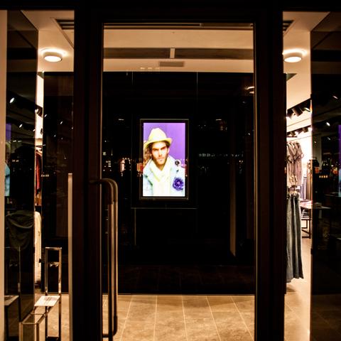 Ein Schaufenster in der Nacht. Von einem Monitor an einer schwarzen Stellwand schaut ein männliches Model mit weißem Hut und weißem Oberteil nach draußen Richtung Betrachter:in. Es steht vor einem violetten Hintergrund.